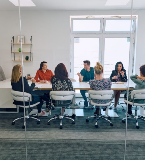 A diverse team of business professionals engaged in a discussion about projects in a modern glass walled office.