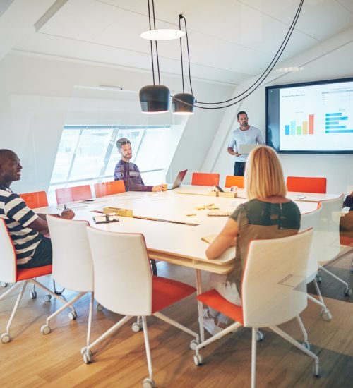 Diverse group of work colleagues and their manager discussing graphs on a monitor while meeting together inside of a glass office